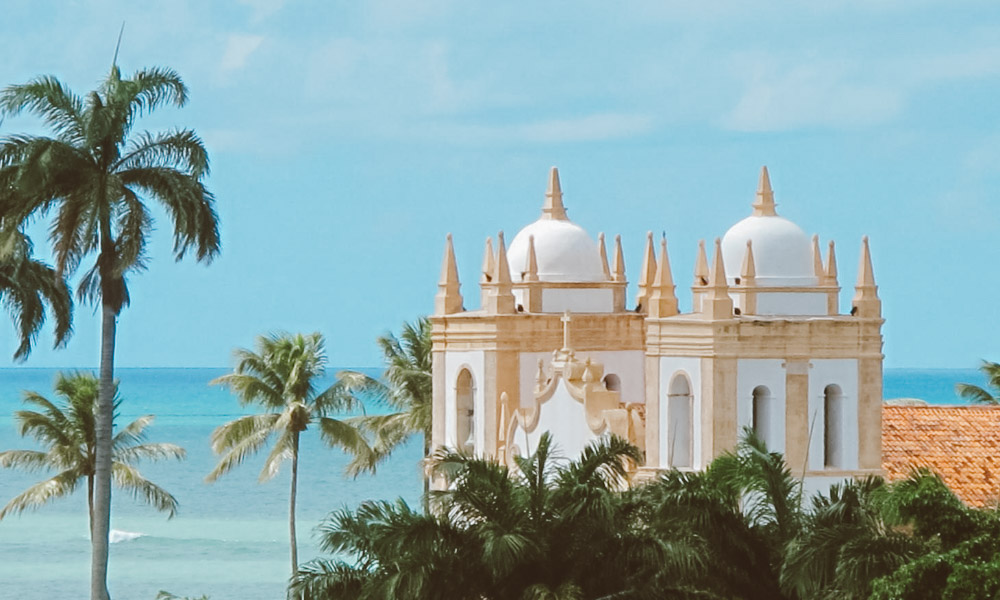 Igreja antiga de frente para o mar, cercada por árvores e céu azul